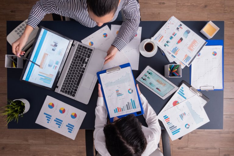An overhead view of two colleagues at a desk comparing printed performance charts, a clipboard and a laptop dashboard, working out which work to keep in house