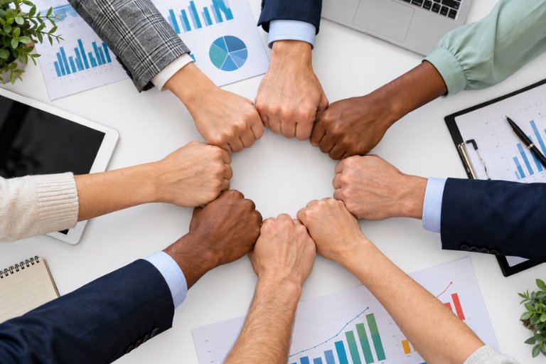 An overhead view of eight colleagues joining fists in a circle above colorful printed charts, a combined internal and partner team growing together