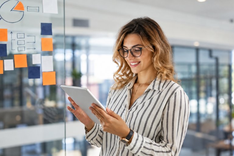 A smiling professional reviewing results on a tablet beside a glass wall of sticky notes, a pie chart and a workflow diagram, weighing the full path instead of a single touchpoint