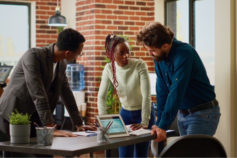 Three colleagues gathered around a table in a brick-walled office, reviewing campaign results together on a tablet and printed notes