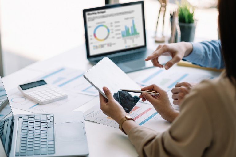 Two colleagues at a desk reviewing a monthly budget dashboard, printed charts and a calculator, weighing campaign results against wider business goals